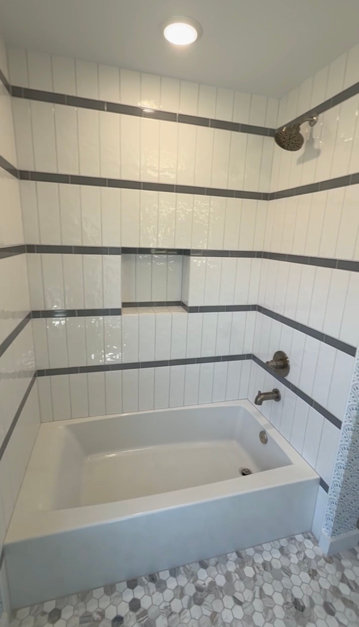 Remodeled tub and shower with white vertical tile, gray accent stripes, and marble hex floor