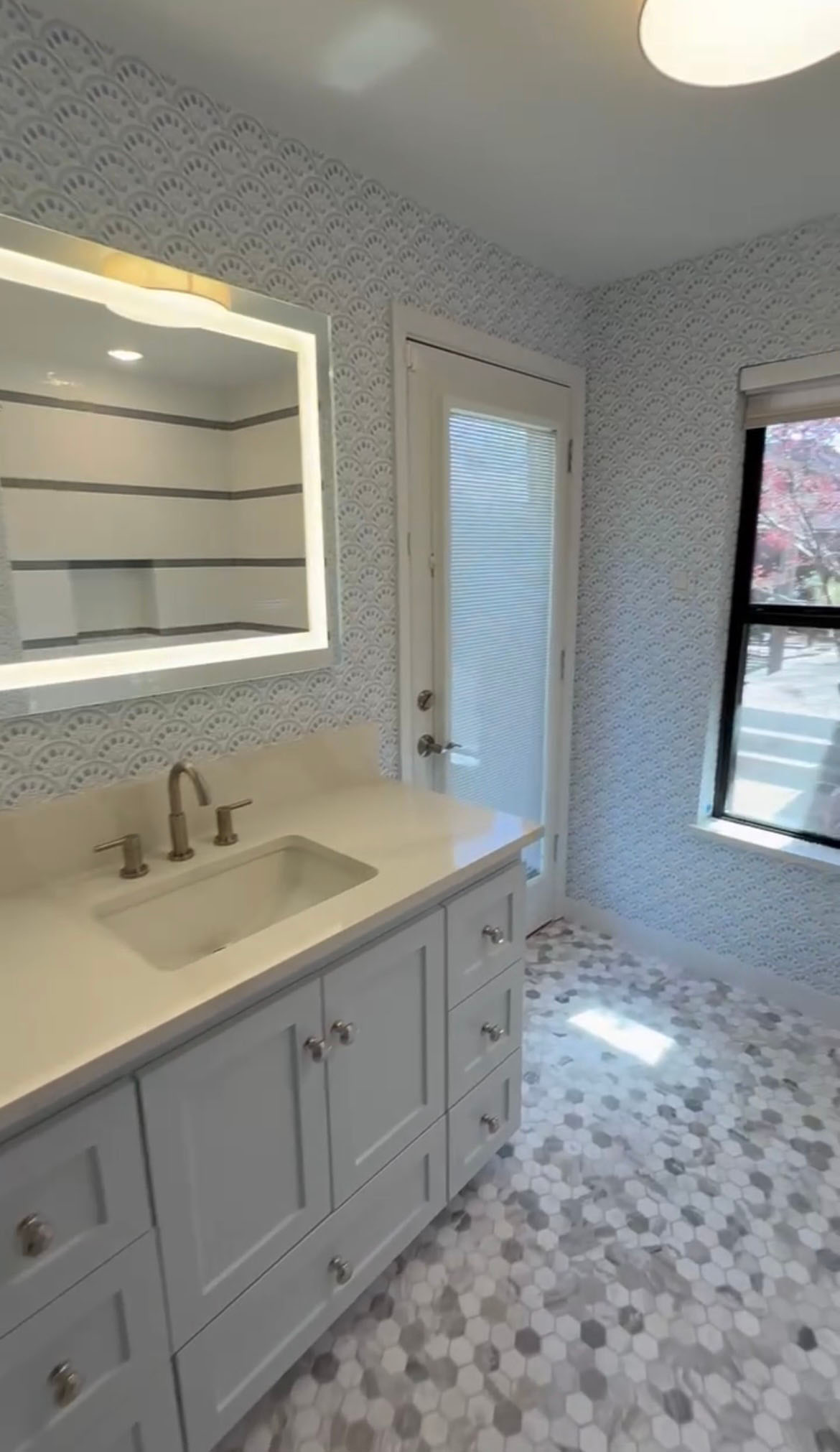 Remodeled bathroom with white vanity, lighted mirror, patterned wallpaper, and marble hex tile floor